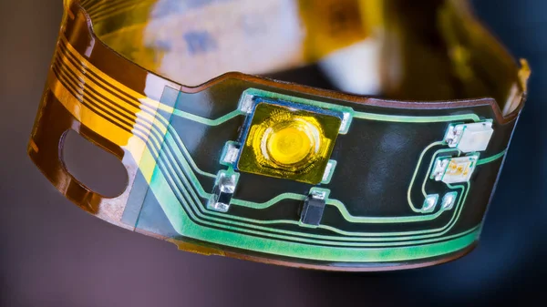 Flex printed circuit and small electronic components in plastic strip curled into circle on a dark background. Closeup of ribbon cable with green and yellow copper lines on flexible PCB from headphones.
