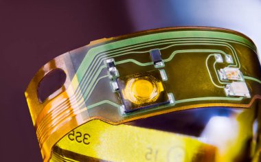 Plastik şeritteki elektronik, esnek baskılı devre mor arka planda halka şeklinde kıvrılmış. Parçalanmış kulaklıklardan gelen yeşil ve sarı çizgiler ve küçük elektrik bileşenleri..