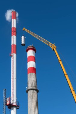 Uzun, yeni ve yenilenmiş bir fabrika bacası ve mavi gökyüzü arka planında vinç patlaması. Havalandırma makinesinin sarı kiriş kolu tamir sırasında bacayı endüstriyel beton bacaya yerleştiriyor..