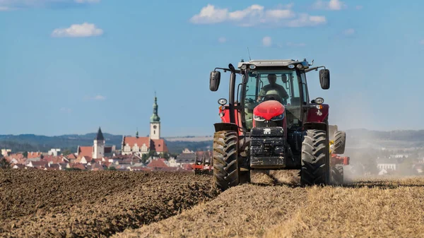 チェコの風景の中で頑丈な泥を耕し 耕す現場の赤いトラクター チェコ市内の教会の塔や歴史的中心部を望む耕地で働く農場車両夏の空の下でタブー — ストック写真