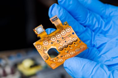 Mühendis elinin mavi eldiveni üzerine basılmış, esnek bir tuş takımı devresi. Yuvarlak düğme düğmeleri, LED diyotlar veya esnek düz kablolar, koyu bulanık arka planda bükülmüş plastik panoda bağlanıcı ile.