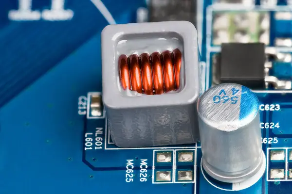 Close-up of electronic coil and electrolytic capacitor on background of blue printed circuit board. Inductor copper wire winding embedded by epoxy in gray case and electrical components on PCB detail.