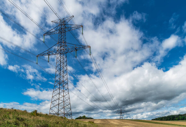 Electric power transmission line in nature scenery with blue sky and beautiful white clouds. Lattice towers and overhead wires to transport electricity fading into distance in stubble of summer field.