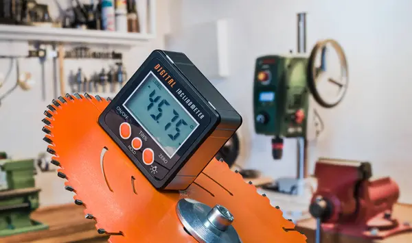 stock image Electronic inclinometer with digital display on orange disc of power miter drop saw. Closeup of precise tool for measuring slope angle and bench vice, drilling machine or lathe on workroom background.