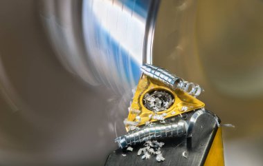 Torna tezgahının metal talaşlı kısmı dönen bir iş parçasına dönüşürken kıvrılarak kıvrılmış. Keskin bıçak kesici ve karbit ekleme ve soyut hareket detayı ile sarmalanmış arkaplan bulanık.