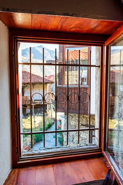 stock image A view through a traditional wooden-framed window in Elati, Trikala, Greece, showcasing the village's stone houses and tiled rooftops with mountainous scenery.