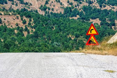 Yunanistan 'ın Kastoria kentindeki ormanlık tepelerle çevrili kırsal bir yolda keskin bir dönüş ve dik bir inişi gösteren yol işaretleri.