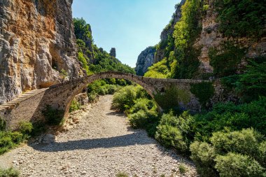 Noutsos (Kokkoris) Bridge, an ancient stone arch bridge in Zagori, Greece, set against a dramatic rocky gorge and lush vegetation. clipart