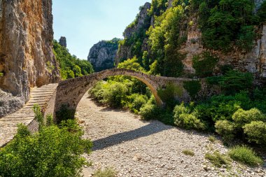 Noutsos (Kokkoris) Bridge, an ancient stone arch bridge in Zagori, Greece, set against a dramatic rocky gorge and lush vegetation. clipart