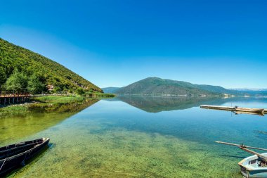 Calm waters of Lake Prespa at Mikrolimni village, reflecting the surrounding mountains with small docks and boats in view. clipart