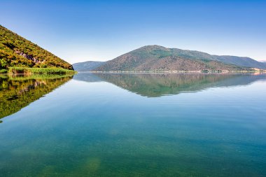 Clear reflections of lush green mountains on the tranquil waters of Lake Prespa under a bright blue sky in Mikrolimni. clipart