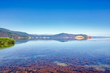 Mikrolimni 'deki açık mavi gökyüzünün altında Prespa Gölü' nün sakin sularındaki yemyeşil dağların berrak yansımaları..