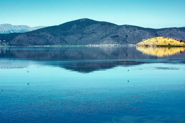 Clear reflections of lush green mountains on the tranquil waters of Lake Prespa under a bright blue sky in Mikrolimni. clipart