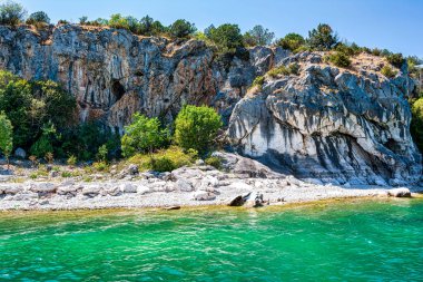 The rocky shoreline of Megali Prespa Lake in Greece, featuring natural cliffs and ancient religious rock paintings surrounded by greenery. clipart