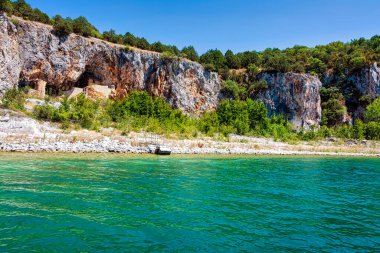 An ascetic building nestled into the rocky cliffs along the shore of Megali Prespa Lake, surrounded by clear waters and natural greenery. clipart