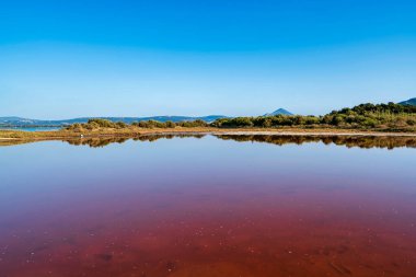 Parlak, sakin bir Gialova gölü manzarası, kırmızı suyla, canlı mavi gökyüzünün altındaki bitki örtüsünü ve dağları yansıtıyor..