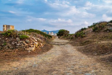 Tarihi Methoni Kalesi 'nin yıkıntılarını kesen kirli bir yol. Etrafı bitki örtüsü ve bulutlu bir gökyüzünün altındaki engebeli araziyle çevrili..