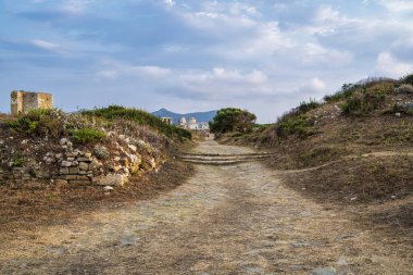 Tarihi Methoni Kalesi 'nin yıkıntılarını kesen kirli bir yol. Etrafı bitki örtüsü ve bulutlu bir gökyüzünün altındaki engebeli araziyle çevrili..