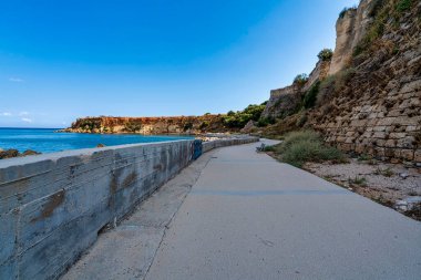 Koroni 'de sakin bir sahil geçidi, taş duvarlarla kaplı ve açık gökyüzünün altında deniz ve kayalık uçurumlar manzarası sunuyor..