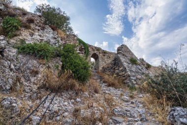 Parçalı bulutlu bir gökyüzüne karşı kurulmuş engebeli kayalık arazi ve aşırı gelişmiş bitki örtüsüyle çevrili tarihi bir taş kemer yolu manzarası.