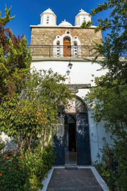 The entrance of Iera Moni Timiou Prodromou features ornate iron gates, lush greenery, and twin white bell towers under a clear blue sky. clipart