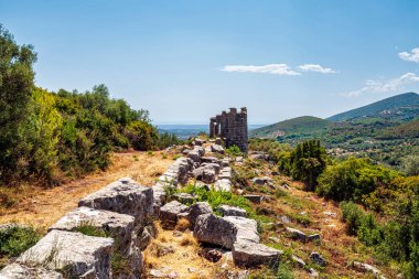 Stone ruins of the ancient walls of Messene, Greece, surrounded by lush greenery and rolling hills under a clear sky. clipart