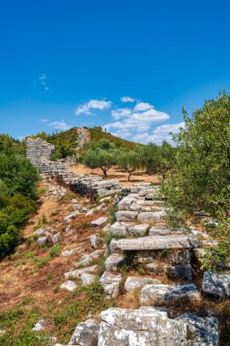 Stone ruins of the ancient walls of Messene, Greece, surrounded by lush greenery and rolling hills under a clear sky. clipart