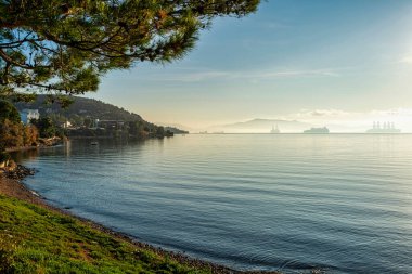 A peaceful misty morning in Elefsina Bay, Greece, with calm waters, silhouetted ships in the distance, and sunlight reflecting on the sea. clipart