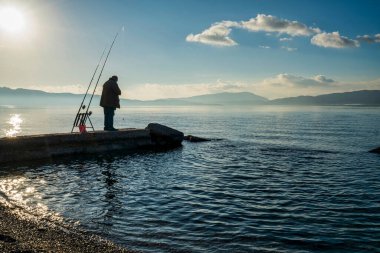 A solitary fisherman stands on a concrete pier near Elefsina Bay, Greece, casting his line into the calm sea under a bright morning sky. clipart