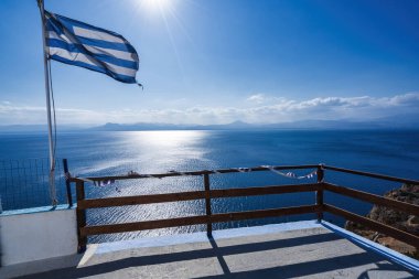 A Greek flag waves in the wind above a coastal viewpoint, overlooking the shimmering blue waters of the Corinthian Gulf under the sun. clipart