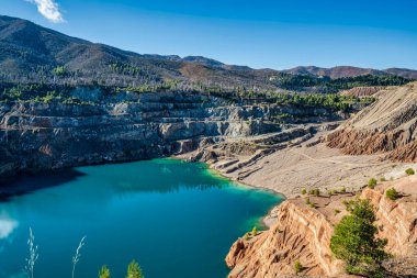 Yunanistan 'ın Evia kentindeki terk edilmiş bir taş ocağında bulunan derin mavi bir yapay göl, katmanlı kaya oluşumları, engebeli arazi ve ormanlı tepelerle çevrilidir..