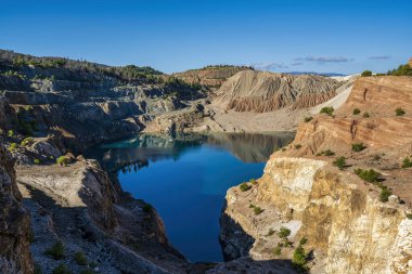 Yunanistan 'ın Evia kentindeki terk edilmiş bir taş ocağında bulunan derin mavi bir yapay göl, katmanlı kaya oluşumları, engebeli arazi ve ormanlı tepelerle çevrilidir..