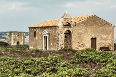 Yunanistan 'ın Methoni kentindeki antik kale kalıntıları yakınında çan kulesi olan yıpranmış taş şapel, kıyı manzarası ve yabani bitki örtüsüyle çevrili..