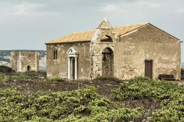 Yunanistan 'ın Methoni kentindeki antik kale kalıntıları yakınında çan kulesi olan yıpranmış taş şapel, kıyı manzarası ve yabani bitki örtüsüyle çevrili..