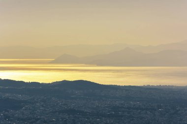Hymettus dağından Atina şehrini parıldayan deniz ve ılık günbatımı ışığı altında uzak adaların siluetleriyle gösteren manzara.