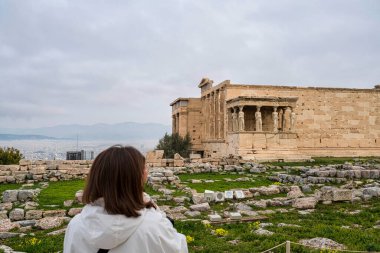 Atina, Yunanistan - 2 Mart 2025: Bir kadın, Akropolis 'teki Echtheion tapınağının tarihi kalıntılar ve bulutlu gökyüzüyle çevrili ünlü Caryatid heykellerini izliyor.