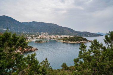 Methana yarımadasının panoramik manzarası, küçük bir liman, kayalık kıyı şeridi ve engebeli dağların desteklediği yemyeşil tepeleri gösteriyor..