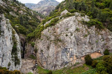 Dik gri kireçtaşı duvarlar dar Aziz Luke Vadisi 'ni, çağlayan akarsuları ve ön plandaki kır çiçeklerinin çerçevelediği gür Akdeniz çam ağaçlarını ortaya çıkarmak için ikiye bölündü..