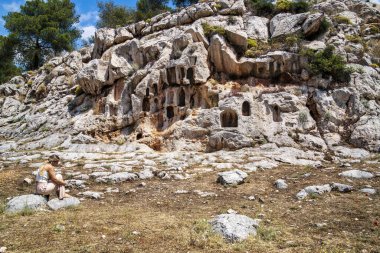 Skaramagas, Yunanistan - 11 Mayıs 2025: Yunanistan 'ın Skaramagas kenti yakınlarındaki Antik Afrodit Sığınağı, adak adakları için kaya oyukları ve sessiz bir tefekkür içinde tek başına oturan bir kadın.