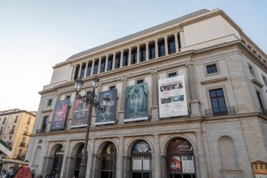 Madrid, İspanya. Teatro Real 'in doğu cephesi Plaza de Isabel II' ye bakıyor. Güneşli bir günde neoklasik binadan sarkan güncel ve yaklaşan opera prodüksiyonlarının reklamını yapan posterlerle.