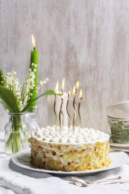 Badem yaprakları ve mumlarla süslenmiş ince katmanlardan yapılmış ekşi krema ve ballı pasta. Kutlama pastası, bir fincan nane çayı, bir buket vadi zambağı ve arka planda mumlar. Kırsal biçim.