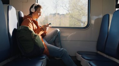 Young man travels alone by electric train with backpack. Man in white wireless headphones scrolls social network pages on smartphone side view