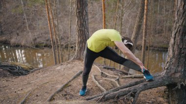 Man in headphones makes stretches standing on tree root near narrow river. Runner in yellow t-shirt warms up before jogging in forest slow motion