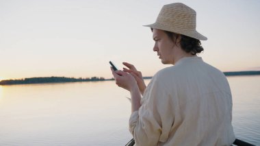 Man tries to get cell signal on smartphone sitting against lake. Guy wearing straw hat sails on boat on large calm forest lake at sunset