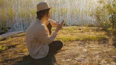 Man tries to get cell signal on smartphone sitting on river bank against reeds at sunset. Guy in straw hat needs connection to call friends