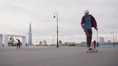 Young man in helmet jumps and makes tricks on skateboard in city on cloudy day. Adventurous guy enjoys riding board on asphalt road slow motion. Russia, St. Petersburg, August 2022