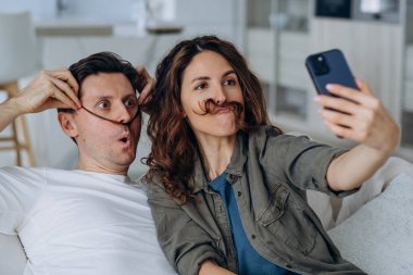 Cheerful in love couple taking a selfie on the phone while sitting on the couch together, making funny mustaches out of their hair and taking pictures