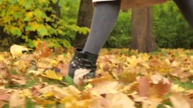 Side view of female legs in black leather boots walking on yellow autumn maple leaves in the park in slow motion. A woman enjoys a walk in nature.