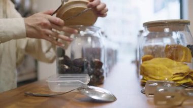 Woman takes dried plums out of glass jar to put into plastic box on wooden table. Female customer buys delicious snacks in supermarket closeup