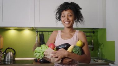 African American woman holds fresh vegetables in market paper bag standing in kitchen. Young housewife with kinky hair looks smiling